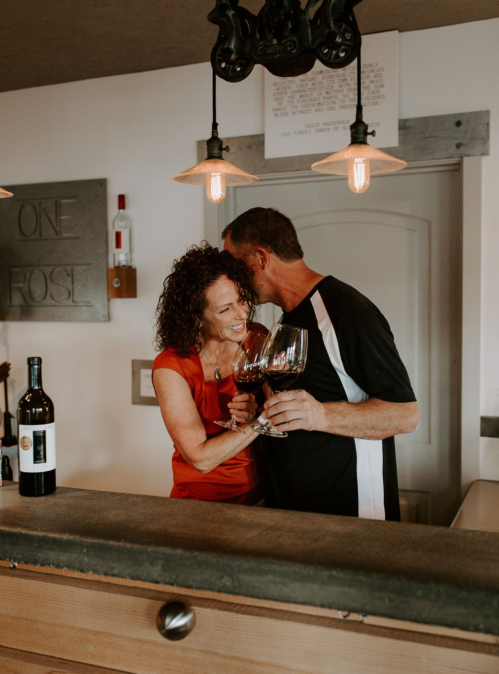 A couple joyfully clinks wine glasses at a cozy bar, surrounded by warm lighting and wine bottles.