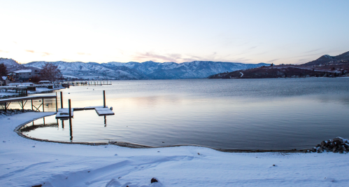 A serene winter landscape featuring a snowy shoreline, calm water, and distant mountains under a clear sky.