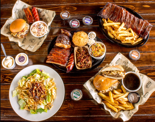 A spread of barbecue dishes including ribs, burgers, fries, coleslaw, and salads on a wooden table.