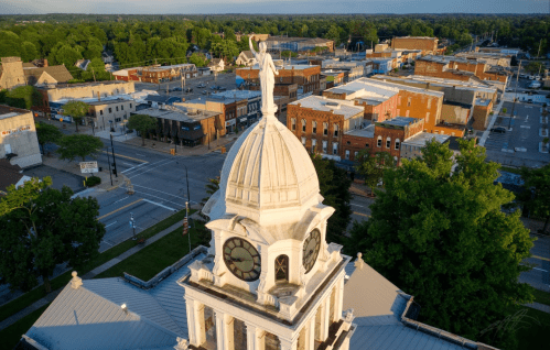 Aerial view of a town with a clock tower and statue on top, surrounded by buildings and greenery.