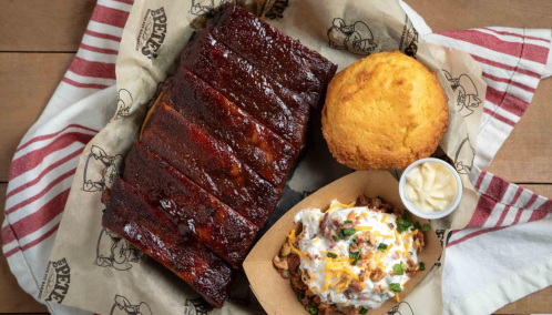 A plate featuring glazed ribs, cornbread, and a loaded baked potato with toppings, served on a checkered paper.