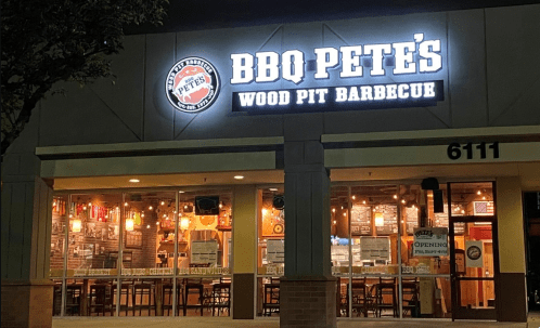 Exterior of BBQ Pete's Wood Pit Barbecue restaurant, featuring a lit sign and large windows showcasing the interior.