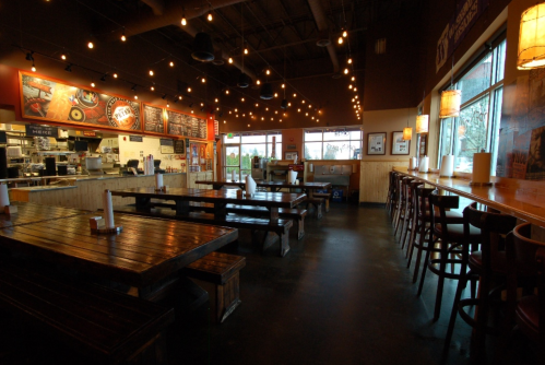Interior of a restaurant with wooden tables, string lights, and large windows, creating a warm, inviting atmosphere.