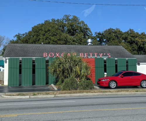 A red car parked outside a restaurant with green shutters and a sign that reads "Boxcar Betty's."