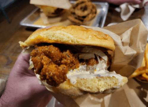 A hand holding a spicy chicken sandwich with crispy chicken, sauce, and slaw on a soft bun, wrapped in paper.