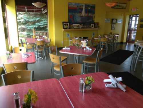 Brightly lit restaurant interior with yellow walls, red tables, and simple wooden chairs, featuring small flower arrangements.