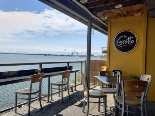 A waterfront patio with empty chairs and tables, featuring a sign for "The Grotto" against a sunny sky.