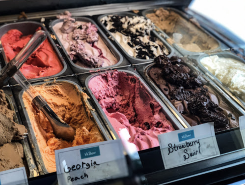 A variety of colorful ice cream flavors in metal tubs, labeled with names like "Georgia Peach" and "Strawberry Swirl."