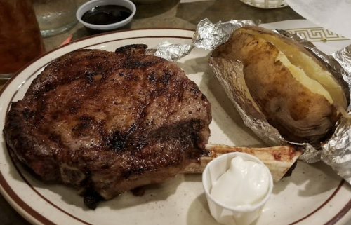 A juicy ribeye steak with a baked potato wrapped in foil and a side of sour cream on a plate.
