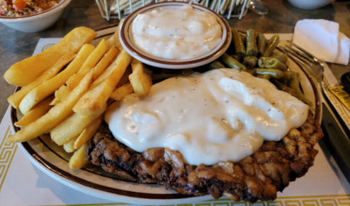 A plate of fried chicken smothered in gravy, served with green beans, fries, and a bowl of mashed potatoes.