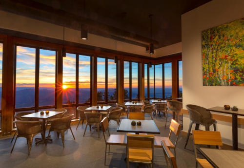 A cozy restaurant with wooden tables and chairs, featuring large windows showcasing a sunset over the mountains.
