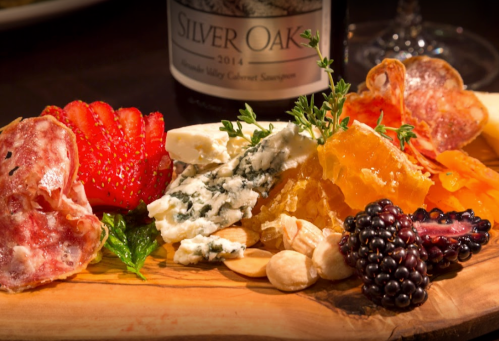 A wooden platter with assorted cheeses, meats, fruits, nuts, and a bottle of Silver Oak Cabernet Sauvignon in the background.