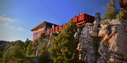 A modern building perched on a rocky cliff, surrounded by trees and bathed in warm sunlight against a blue sky.