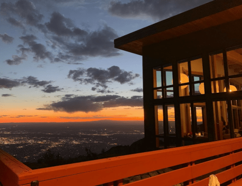 A sunset view from a mountain cabin, featuring a red railing and large windows reflecting the colorful sky.
