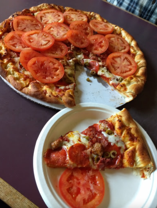 A pizza topped with sliced tomatoes, with one slice on a paper plate beside the whole pizza.