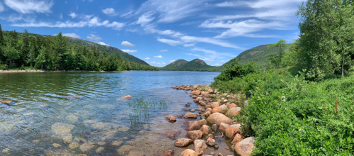 A serene lake surrounded by green hills and rocky shores under a blue sky with fluffy clouds.