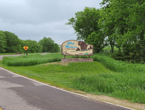 Sign for Blue Mounds State Park, surrounded by green grass and trees, along a winding road on a cloudy day.