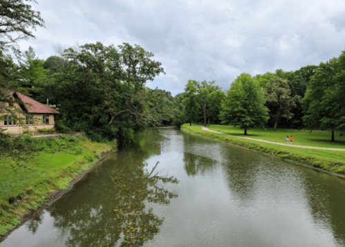 A serene river flows beside a green park, with trees lining the banks and a building in the background under a cloudy sky.