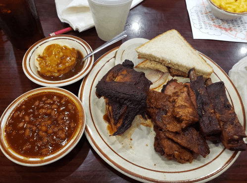 A plate of barbecue featuring ribs, chicken, and sides of baked beans and macaroni and cheese, with bread on the side.