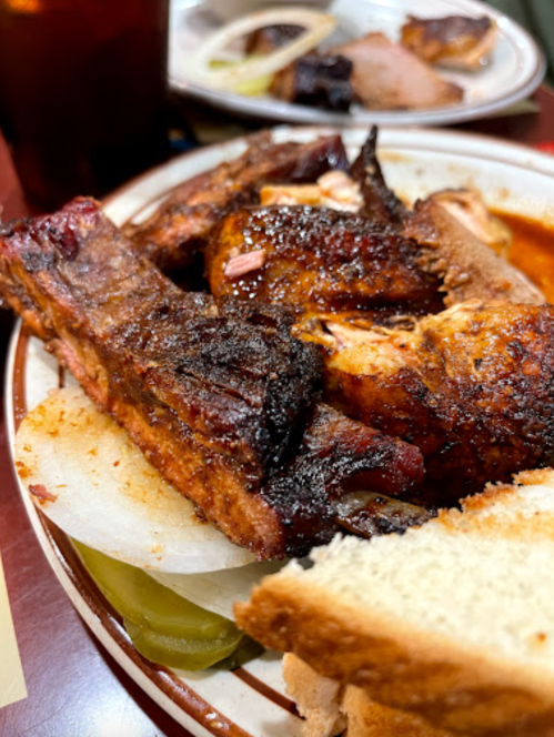 A plate of grilled ribs and chicken, served with pickles, onions, and slices of bread.