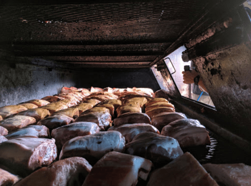 A close-up view of various meats being smoked inside a large smoker, with smoke and heat visible.
