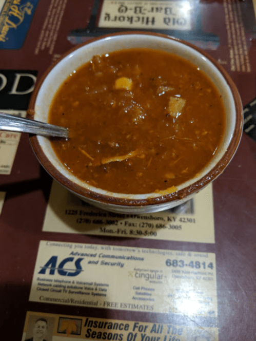 A bowl of thick, brown soup with chunks of meat and vegetables, accompanied by a spoon, on a table with printed text.