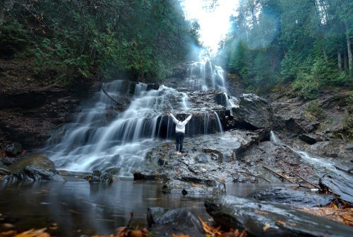 A person stands in front of a cascading waterfall surrounded by lush greenery and misty air.