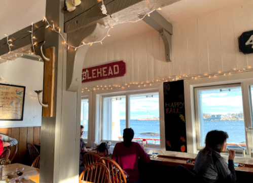 Cozy restaurant interior with string lights, wooden decor, and a view of the water and city skyline through large windows.