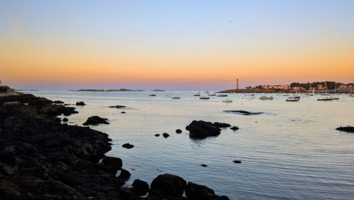 A serene coastal scene at sunset, featuring calm waters, boats, and a distant lighthouse against a colorful sky.