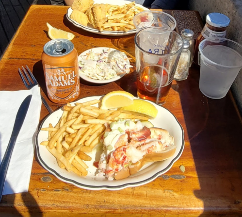 A plate with lobster roll, fries, and lemon, alongside a beer and coleslaw on a wooden table.