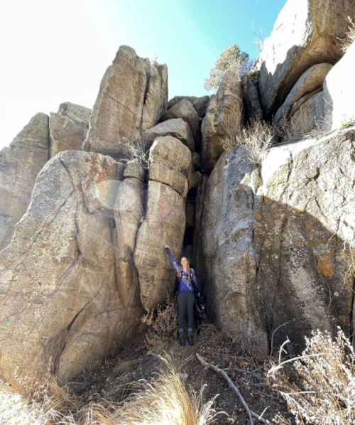 A person stands beside a large rock formation, raising one arm in a natural landscape with dry grass and clear blue sky.