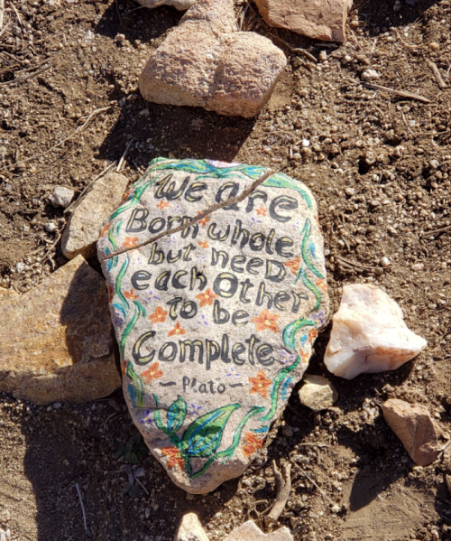 A painted rock with the quote: "We're born whole but need each other to be complete. Plato" surrounded by small stones.