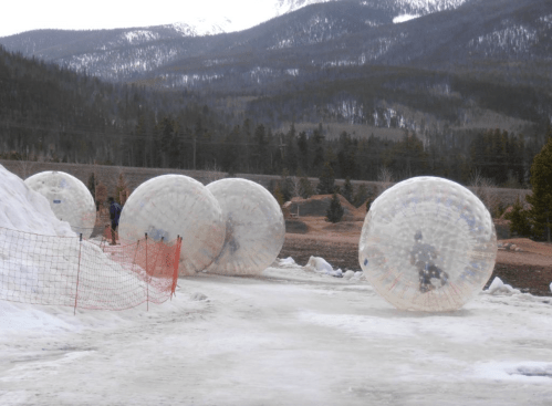People inside transparent inflatable balls roll on a snowy landscape with mountains in the background.
