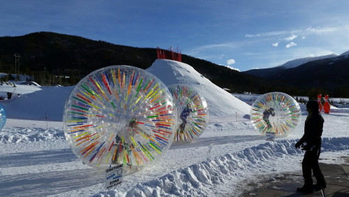Colorful inflatable zorb balls on snow, with a snowy hill and mountains in the background under a clear blue sky.