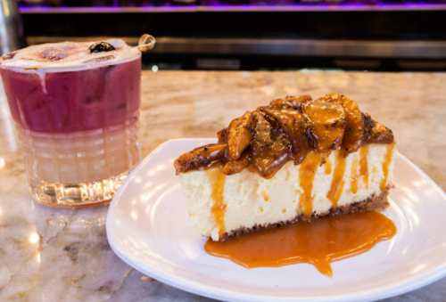 A slice of caramel apple cheesecake drizzled with sauce, alongside a colorful cocktail on a marble countertop.