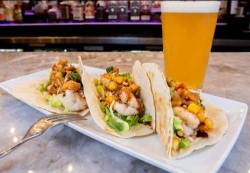 Three shrimp tacos topped with mango salsa and lettuce, served on a white plate with a pint of beer in the background.