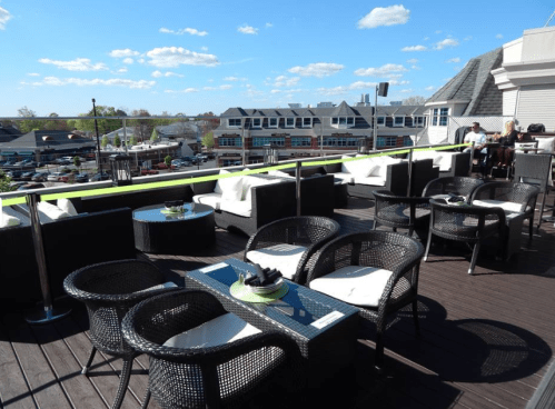 A rooftop patio with wicker furniture, tables, and a view of a bustling area under a blue sky with clouds.