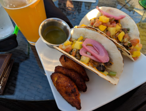 Two tacos topped with mango and pickled onions, served with a side of fried plantains and a drink.