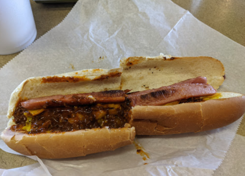A split hot dog bun filled with chili, mustard, and slices of meat, resting on a piece of white paper.