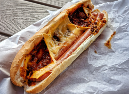 A loaded hot dog in a bun topped with melted cheese and chili, resting on white paper.