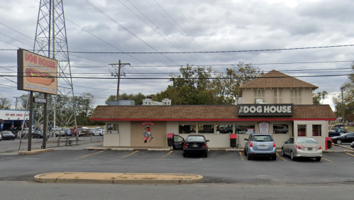 A small restaurant called "The Dog House" with a sign featuring a hot dog, surrounded by a parking lot and power lines.