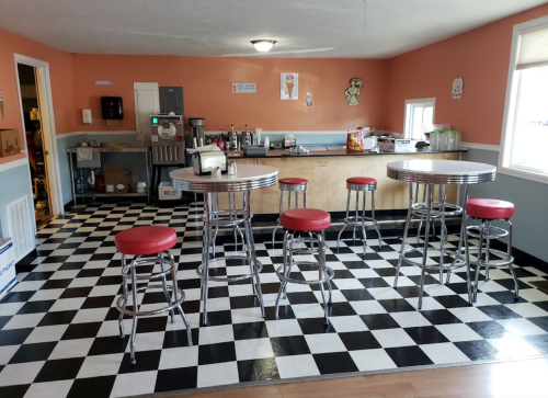 A retro-style diner with checkered floor, chrome tables, red stools, and a counter in a cozy, brightly colored room.