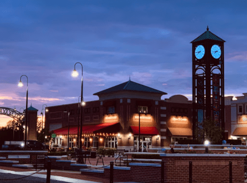 A clock tower stands beside a brick building at dusk, with streetlights illuminating the area and a colorful sky in the background.