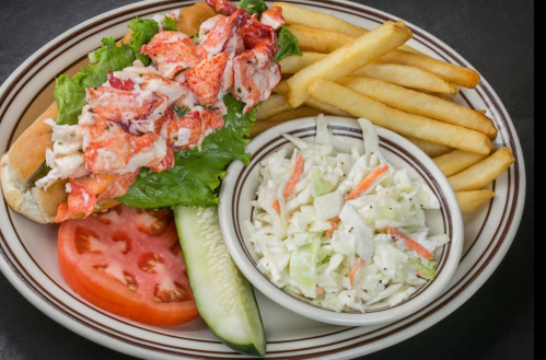 A lobster roll on a bed of lettuce, served with fries, a slice of tomato, cucumber, and a side of coleslaw.