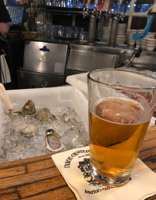 A glass of beer sits on a bar next to a bucket of ice with oysters, with a bartender in the background.