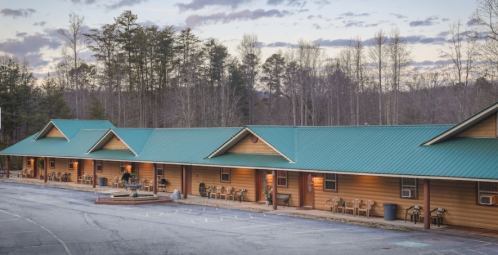 A rustic lodge with a green metal roof, surrounded by trees, featuring outdoor seating and a gravel parking area.