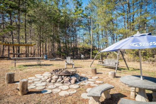 A rustic outdoor seating area with a fire pit, surrounded by trees and picnic tables, under a clear blue sky.