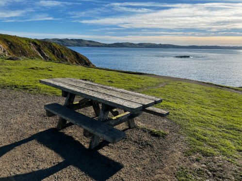 A wooden picnic table on a grassy area overlooking a calm sea and distant hills under a clear blue sky.