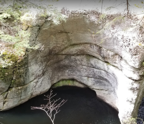 A rocky cliff overhangs a dark pool of water, surrounded by greenery and trees.