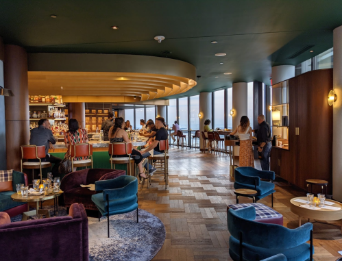 A stylish bar with patrons seated at tables and the counter, featuring modern decor and large windows with a city view.